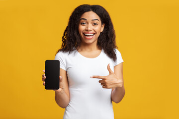 Naklejka premium Positive black lady showing modern smartphone, pointing at empty blank screen, recommending useful mobile application, standing over yellow background, copy space, studio shot