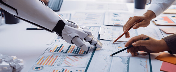 A robotic arm and human hands collaborate on analyzing business data and charts in a modern office, showcasing the fusion of technology and human expertise.Noogenesis