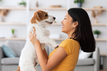 Emotional young asian woman petting her cute dog at home, loving korean lady playing with fluffy...