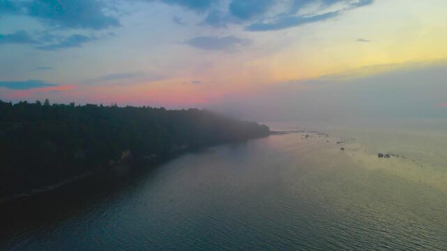 Cinematic aerial reveal of a foggy coastline at sunset with forested cliffs on one side and calm turquoise ocean reflecting warm orange and blue sky tones.