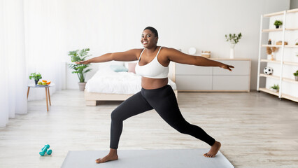 Obraz premium Strength yoga for weight loss. Curvy black lady standing in warrior asana, exercising on sports mat at home, panorama. Young overweight woman doing domestic workout, practicing pilates during covid