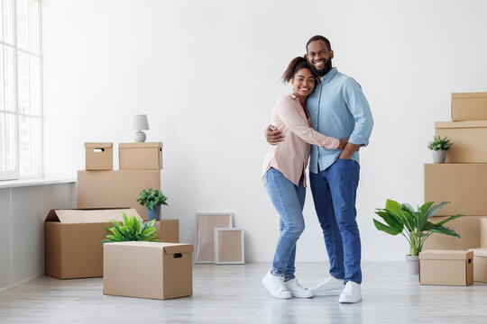 Smiling young african american family hugging in new apartment with boxes and plants in room interior. Married couple enjoying result of renovation, moving in apartment or buying of property, indoor - Powered by Adobe
