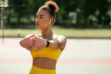 Portrait of confident sporty African American woman in wireless earbuds warming up before jogging...