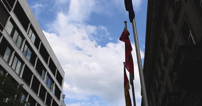 Street view in Frankfurt am Main with modern and historic buildings lining a narrow urban street. Flags waving on flagpoles against a partly cloudy sky, capturing the everyday city atmosphere of a maj