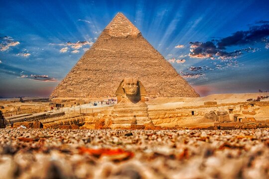 A majestic low-angle view of the Great Sphinx of Giza with the Pyramid of Khafre (Chephren) towering in the background. Bright sunbeams radiate from behind the pyramid, creating a dramatic effect agai