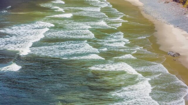 An aerial drone shot captures gentle ocean waves as they roll onto the golden sandy shore of Sand Lake on the Oregon Coast. The white foam creates a rhythmic pattern, enhancing the warm coastal scener