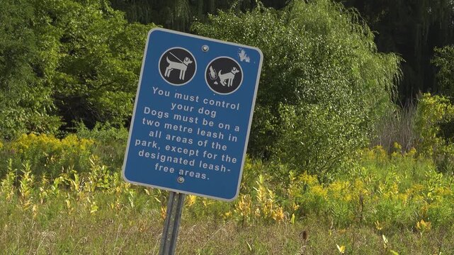 A blue sign with rules about walking dogs in a sunny field and forest at Bronte Provincial Park during the summer.