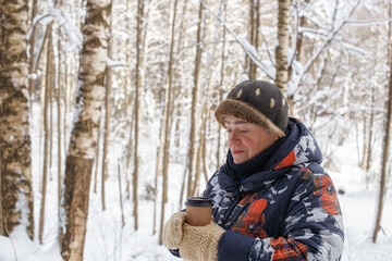 Fototapeta premium A happy elderly woman, with a cup of coffee in her hands, spends her leisure time in the winter park.