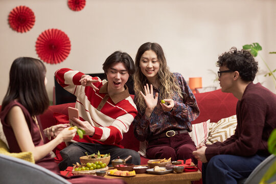 Group of Asian young adults sitting together on sofa celebrating holiday, smiling and posing for selfie with smartphone, sharing food and enjoying festive Tet gathering indoors