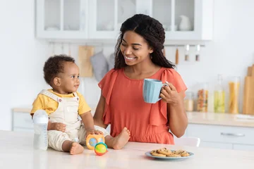 Fotobehang Onderzeeër Portrait Of Happy Black Family Mom And Infant Son Spending Time In Kitchen At Home, Beautiful African American Mother Drinking Tea While Her Cute Toddler Boy Sitting On Counter Table, Free Space  © Prostock-studio