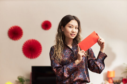 Portrait of young adult Asian woman smiling and holding red envelope with both hands, standing indoors with festive decorations in background during holiday celebration