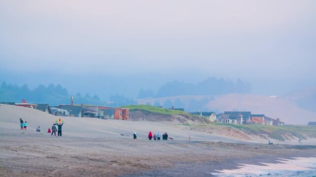 A wide-angle shot of a sandy beach on a misty or foggy day. People are scattered along the shoreline near the water's edge. In the background, a row of colorful buildings sits atop a grassy dune or hi