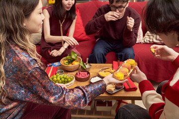 Group of young Asian women and young Caucasian men sitting together sharing traditional holiday food and snacks, celebrating Tet holiday with mooncakes and fruit on table