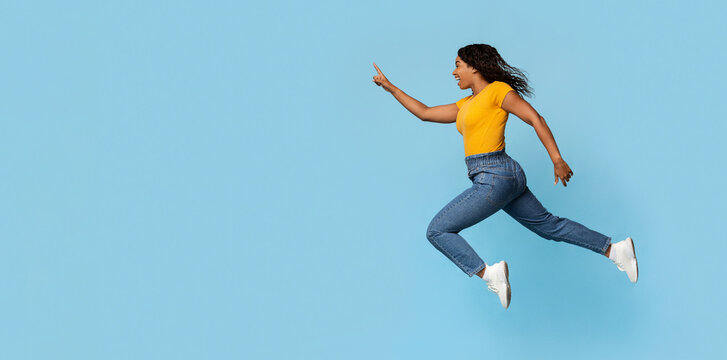 Excited young black pretty lady jumping towards copy space, emotional african american woman running in the air and pointing at advertisement over blue studio background, panorama, web-banner