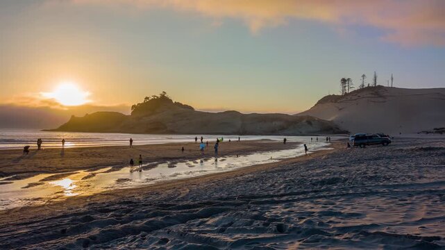People walk along the beach at Cape Kiwanda State Natural Area in Pacific City, Oregon, at sunset. The iconic Haystack Rock formation is visible offshore, with the large sand dune looming in the backg
