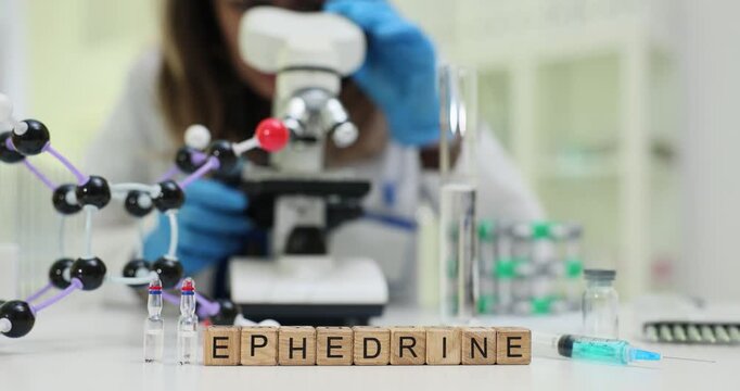Laboratory worker conducts careful analysis with microscope. Miniature wooden cubes on table form word Ephedrine by ampoules syringe and molecular