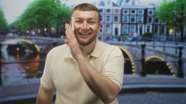 Man pressing palm to cheek on an amsterdam canal street near lit bridge arches and riverside buildings, looking toward camera with a half smile; playful curiosity.