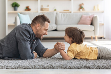 Fototapeta premium Bringing up strong kid. Happy father competing arm wrestling match with his little boy, lying together on floor at home and smiling to each other, profile portrait
