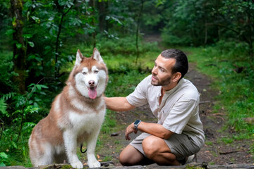 Guy crouches down and plays with his brown and white Siberian Husky in a wooded area. The happy dog gives the man his paw. The dog has a leash attached to its collar.