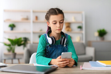 Portrait of serious teenage kid girl holding and using smartphone browsing social network apps, surfing internet and scrolling news feed, reading electronic e-book, sitting at table in living room