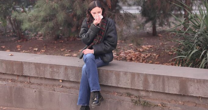 Young woman blows runny nose and yawns sitting on stone parapet slow motion. Brunette female sneezes feeling cold symptoms in autumn city park