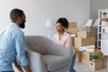 Fototapeta premium Happy millennial african american husband and wife carry armchair in apartment with cardboard boxes. Moving in flat, new home and relocation, positive emotions of owners, buying and renting house