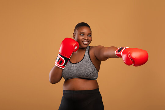 Self-Defence Classes. Portrait Of Young Curvy Black Woman Wearing Boxing Gloves, Confident African American Female Standing Over Brown Background In Studio, Ready To Attack, Making Throwing Punch
