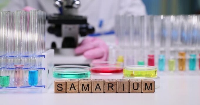 Chemist adjusts microscope controls to improve clarity. Wooden letter cubes on table clearly form word Samarium near test tubes with reagent liquids