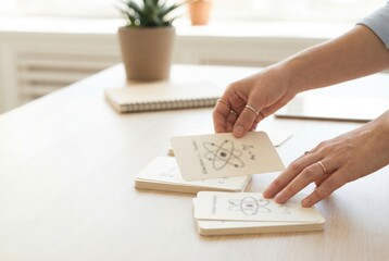 Person studies cards with scientific concepts indoors on a wooden table during daytime with plants and notebooks in the background