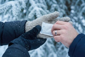 Two people share warmth in snowy weather with hands exchanging a heating pad while standing in a winter landscape surrounded by trees