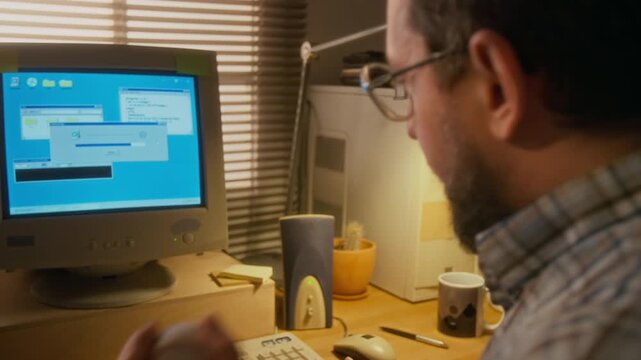 Close-up of middle-aged man in glasses sitting at home at desk, working on vintage computer, waiting for dial-up modem to connect to internet, throwing up and catching baseball