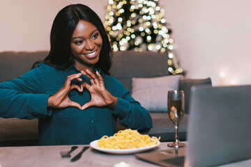 Obraz premium A woman sits at a table enjoying pasta while looking at a laptop. She smiles and makes a heart shape with her hands. A Christmas tree with lights is in the background.