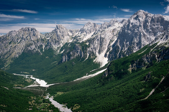 Panoramic summer view of the dramatic limestone peaks of the Accursed Mountains rising steeply above the lush green forests and dry riverbed of Valbona Valley in northern Albania