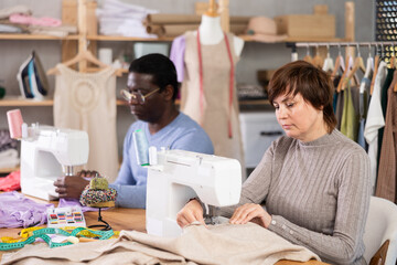 Two tailors, middle-aged man and woman, working side by side at sewing machines in tailoring studio, focusing on assembling fabric pieces with precision and control