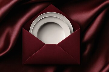 Elegant white dining plates emerging from a deep burgundy envelope invitation on silk