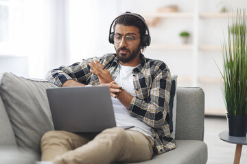 Emotional indian guy sitting on couch at home, having video call with partner, yelling and gesturing at notebook screen, using headset, middle-eastern man fighting with colleagues or lover