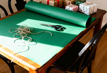 Green wrapping paper roll with scissors and twine on a wooden table. Christmas gift packaging preparation with presents in background. Holiday DIY craft concept with copy space
