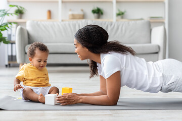 Fototapeta premium Healthy Lifestyle Concept. Young Black Mother Training At Home With Cute Little Baby Playing Around, Beautiful African American Woman Doing Plank Exercise And Looking At Her Adorable Infant Child