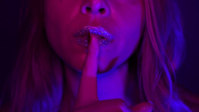 Young blonde woman with glitter lips holding finger to her mouth in silence gesture, sensual beauty close-up under pink and blue neon light. Feminine mysterious mood, elegance, intimacy