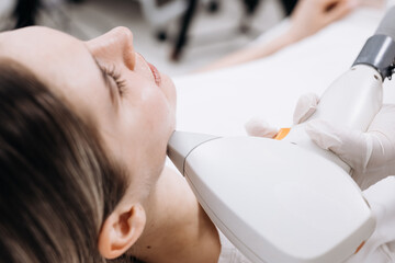 Female patient receiving laser hair removal treatment on her chin with a handheld device in a modern clinic setting, soft lighting and medical equipment visible in the background © wedmoments.stock