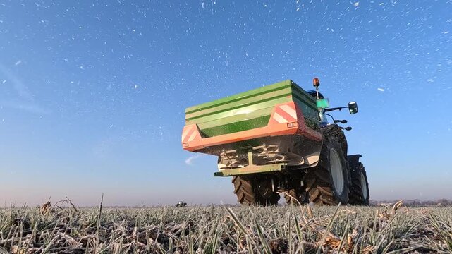 Farmer with agricultural machinery fertilizing wheat field in winter time, slow motion. Spreading mineral fertilizer, landscape rural scene - low angle shot
