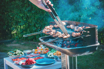 A grill full of meat roasting, hands with tongs turning the meat so it doesn't burn.