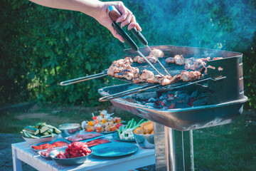 A grill full of meat roasting, hands with tongs turning the meat so it doesn't burn.