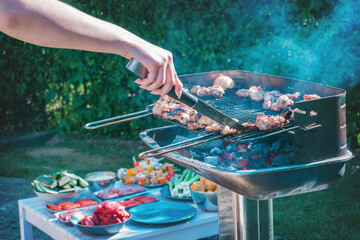 A grill full of meat roasting, hands with tongs turning the meat so it doesn't burn.