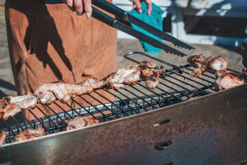 A grill full of meat roasting, hands with tongs turning the meat so it doesn't burn.