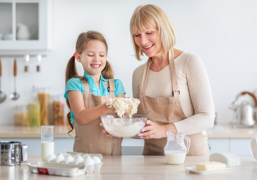 Cooking With Pleasure. Happy little girl in apron mixing dough for cookies or pancakes in glass bowl, cheerful senior grandma helping her grandchild at kitchen. Smiling granny sharing family recipe