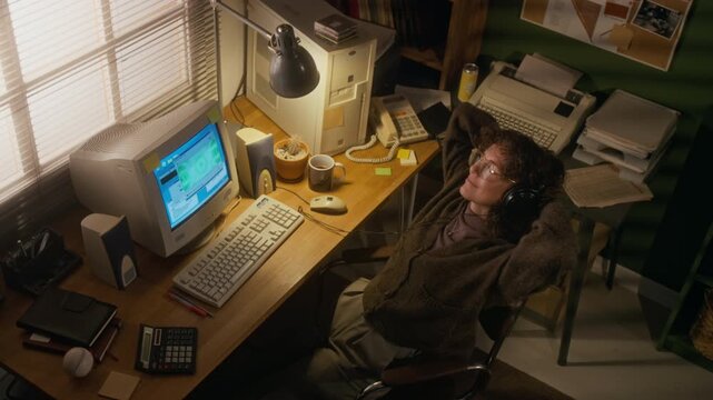 Top view of Caucasian woman in brown woolly cardigan and glasses, resting in chair while working on vintage computer in home office, listening to music, next to retro corded telephone and fax machine