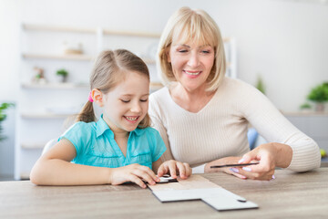Fototapeta premium Loving smiling mature woman sitting at table with little girl engaged in game assemble jigsaw together, happy grandmother playing having fun with preschooler kid making picture connect puzzle pieces