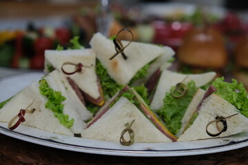 A stack of crustless triangle sandwiches, filled with ham, cheese, and fresh lettuce, is held together with decorative bamboo picks on a white serving plate in a dining setting.