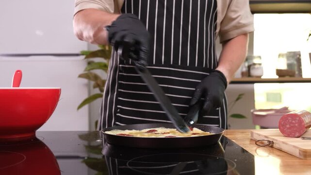 Man in apron pours broken liquid eggs into frying pan with sausage slices in kitchen time-lapse. Male chef cooks nutritious breakfast at home